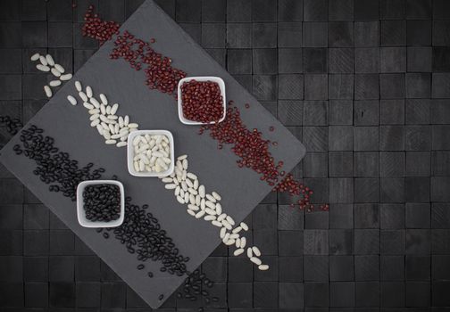 Background design composition from above with square slate plate and three bowls with beans of different colors that make up three colored stripes white, red and black on a black background