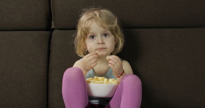 Girl sitting on sofa and eating corn puffs. Child taste puffcorns
