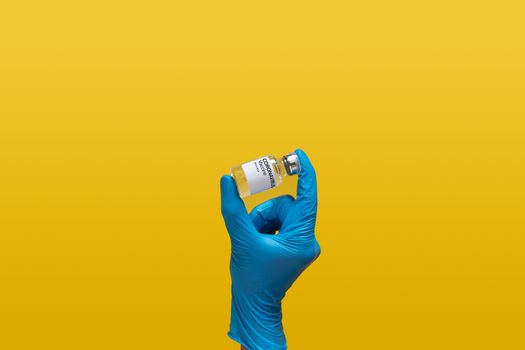 doctor at laboratory wearing blue rubber glove holding Coronavirus vaccine dose bottle label, isolated on yellow background. laboratory analyzing for testing and invent drug and vaccine for covid-19