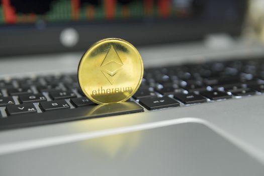 Golden coin of Ethereum on a black keyboard of silver laptop and diagram chart graph on a screen as a background. Virtual cryptocurrency concept. Mining of ethereums online bussiness. Trading.
