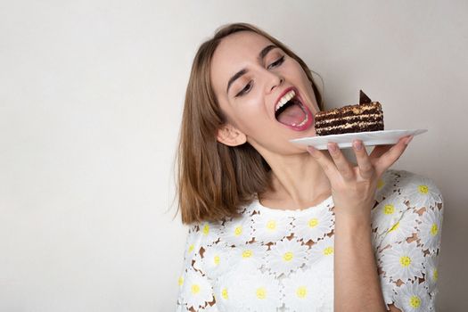 Pleased woman bites tasty chocolate cake over a grey background. Empty space