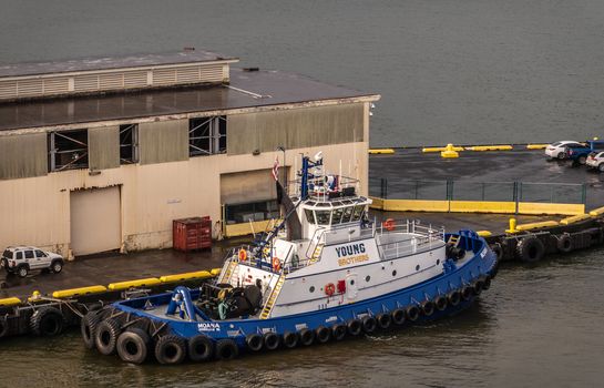 Hilo, Hawaii, USA. - January 14, 2020: Closeup of Young Brothers blue-white tugboat at quay with beige warehouse under rain. Dark ocean water.