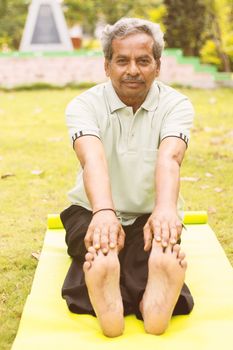 Low-angle full length view of a old man sitting down on exercise or yoga mat touching his toes - Concept of active happy elderly health and fitness