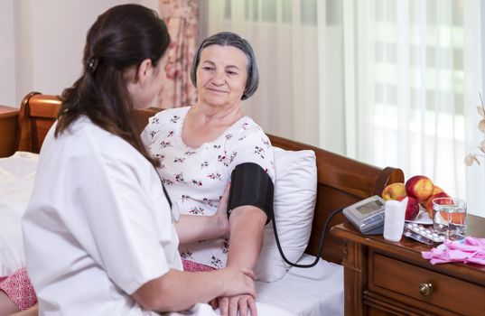 Female doctor checking blood pressure to senior woman at home or in nursing home. Elderly healthcare concept.