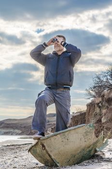 man stands in a broken boat on the beach and carefully looks through binoculars