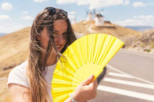 Blonde girl with a yellow fan in her hand fanning herself. Traditional windmills in the background.