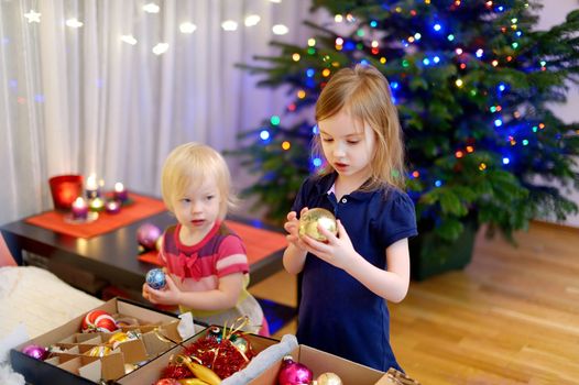 Two adorable little sisters decorating a Christmas tree with colorful glass baubles at home