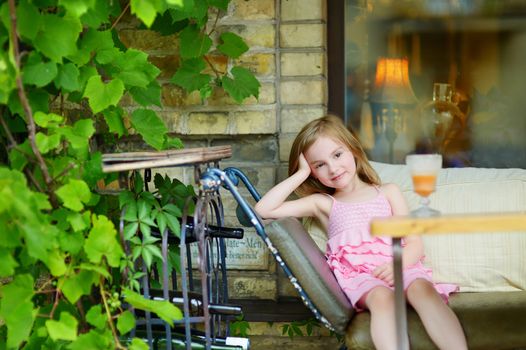 Adorable little girl drinking orange juice in an outdoor cafe on warm and sunny summer day