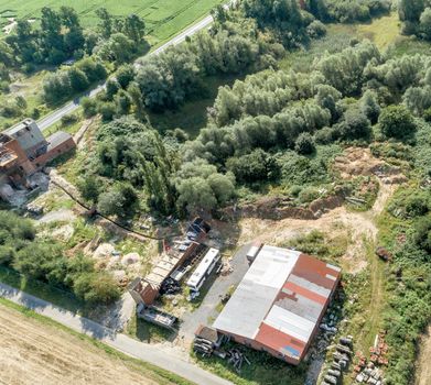 Downsized plant for the repair of machines with a scrap yard behind a forest with a wild dump, diagonally shot aerial photograph.