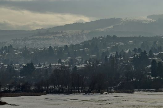 Winter scene with frozen lake, snowy mountain, glade, forest and residential district of bulgarian village Pancharevo