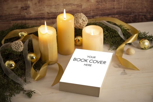 Christmas setting for a book presentation: book with blank cover settled with three lit candles , white organza and gold satin ribbons, gold baubles on light wooden table and dark wood background