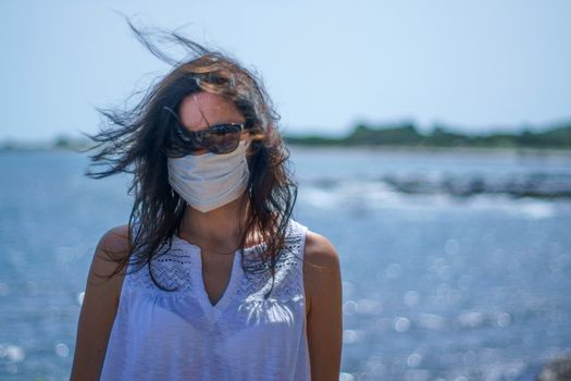 Coronavirus seaside holidays: half-length shot of a woman at the beach with the mask for Covid-19 pandemic with cloudy sky