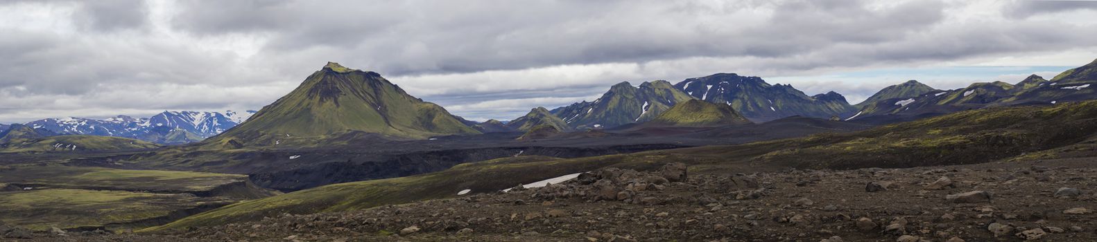 Colorful wide panorama, panoramic view on volcanic landscape in Nature reserve Fjallabak in central Iceland with green Maelifell hill and blue snow covered mountain range, moody sky background