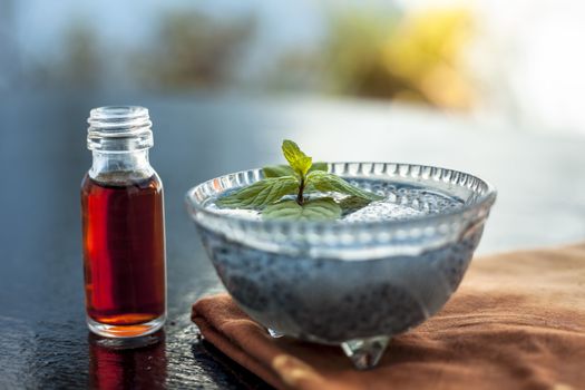 Glass bowl full of soaked sabja seeds or falooda seeds or sweet basil seeds with its extracted essence or essential oil in a transparent glass bottle on wooden surface.