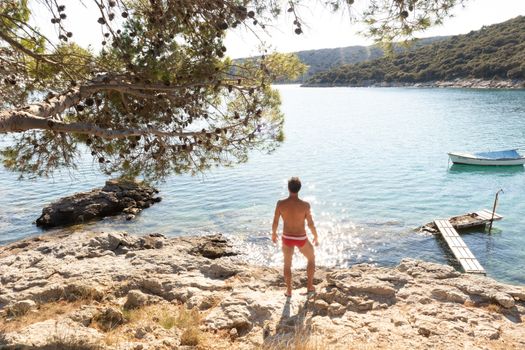 Rear view of man wearing red speedos tanning and realaxing on wild cove of Adriatic sea on a beach in shade of pine tree.