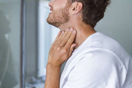 Thyroid self-exam checkup. Young man touching his neck at home bathroom doing self-check of his thyroid gland looking at mirror for early signs of health problem