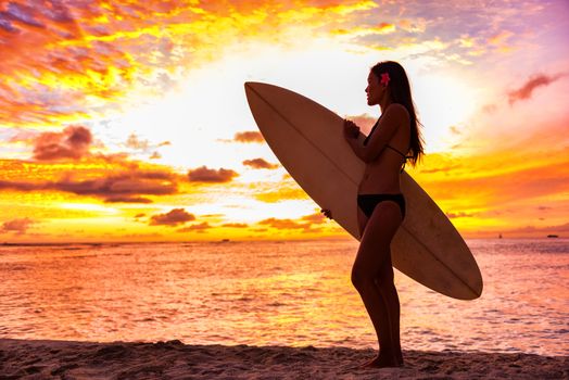 Surfer bikini girl on Hawaii beach holding surf board watching ocean waves at sunset. Silhouette of Asian sport woman over landscape, sky and clouds background. Summer vacation lifestyle
