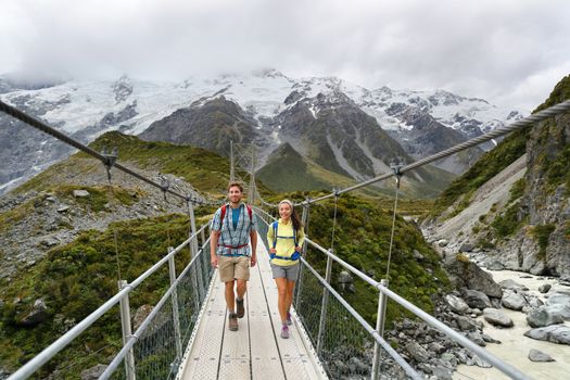 Tourists walking crossing bridge on the Hooker Valley Track hiking trail, New Zealand. Hikers people at Aoraki, Mt Cook National Park with snow capped mountains landscape