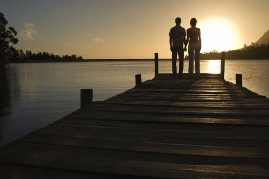 Couple standing on dock by lake holding hands back view.