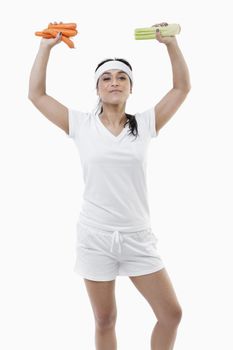 Portrait of young sportswoman with arms raised holding carrots and celery over white background