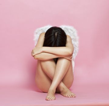 Sad Asian woman with angel wings sitting on floor against pink background