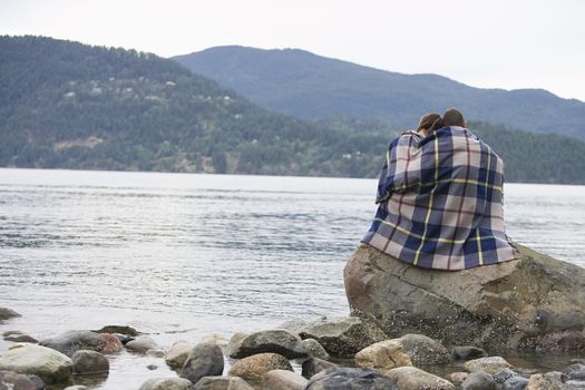 Couple wrapping in blanket sitting on rock by ocean