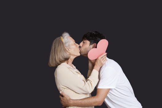 Young man kissing senior woman with red paper heart against black background