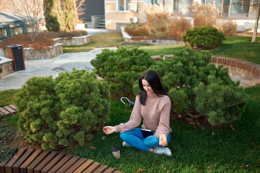 Charming lady practicing meditation on a green lawn outside of building