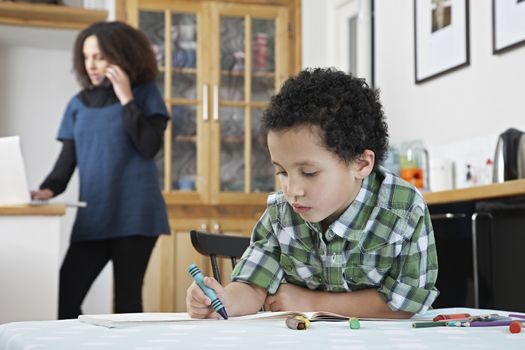 Boy colouring with crayon mother on phone in background