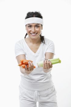 Portrait of young sportswoman holding carrots and celery over white background