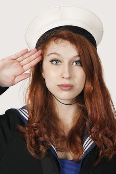 Portrait of young woman in sailor's uniform saluting against gray background