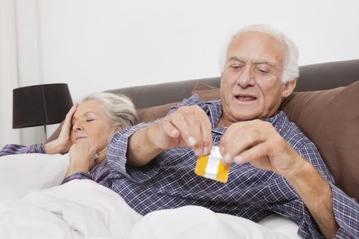 Elderly man tearing condom packet while spouse sleeping behind on bed