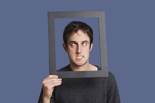 Young man making funny faces through frame over blue background
