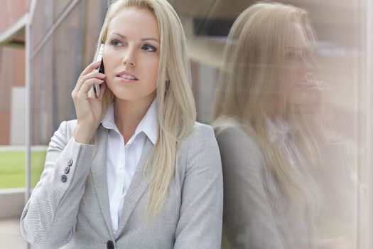 Beautiful blond businesswoman conversing on cell phone while looking away by glass wall