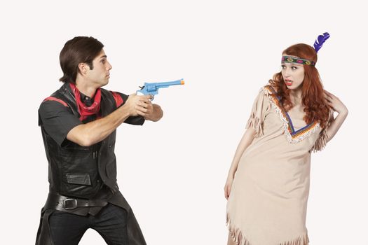 Young cowboy aiming gun at woman in old-fashioned costume against gray background