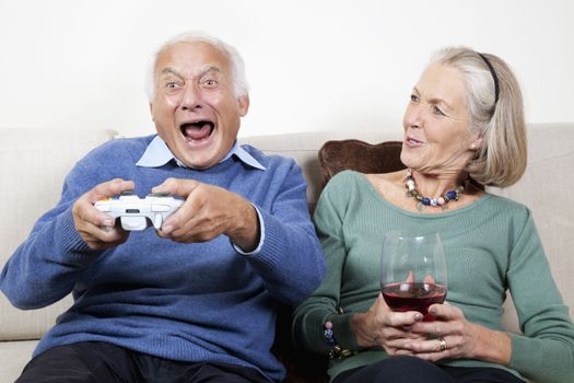 Happy senior woman with wine glass looking at excited spouse playing video game