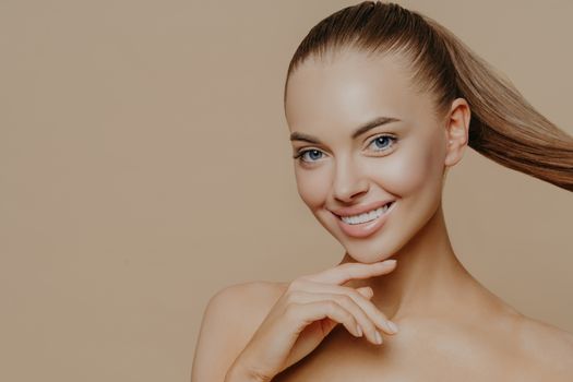 Tranquil undressed woman with healthy perfect skin, dark hair combed in pony tail, enjoys facial treatments, has well cared complexion, stands against beige studio wall, empty space. Skin care concept