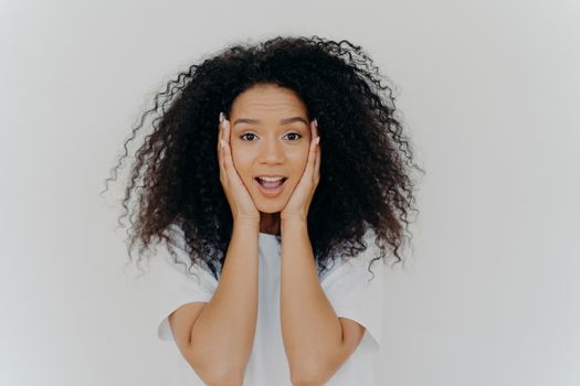 Photo of surprised cheerful woman with Afro haircut, keeps both hands on cheeks, has natural beauty, opens mouth, cannot believe in exciting news, wears t shirt, poses against white background.