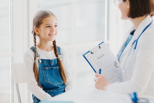 Happy little child listens attentively doctors advice and prescription, comes to see pediatrician in clinic, has two pigtails, wears denim overalls, talk about health problems, has checkup examination