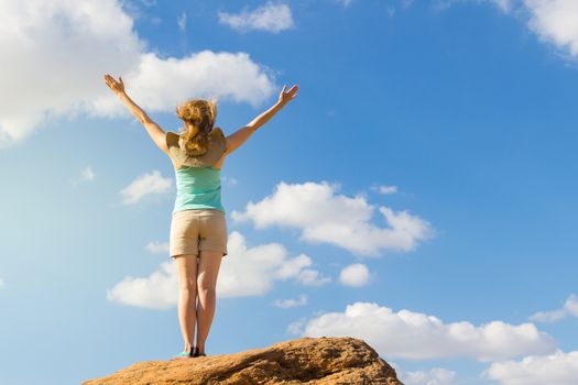 Young woman in shirt and shorts with raised hands is staying on stone. Blue sky with clouds. Success and freedom concept.