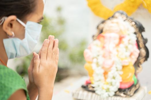 Girl Kid in medical mask praying by closing eyes in front of Lord Ganesha during Ganesha or vinayaka Chaturthi festival - concept of festival celebrations during covid-19 or coronavirus pandemic.