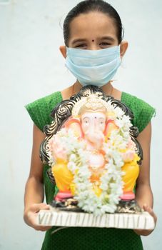 Girl Kid with medical mask holding Ganesha Idol on isolated background - concept of vinayaka Chaturthi festival celebrations during coronavirus or covid-19 pandemic