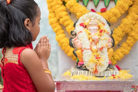Girl Kid praying by closing eyes and folding hands in front of Lord Ganesha Idol during Ganesha or vinayaka Chaturthi festival ceremony at home - concept of Indian religious festival celebrations.
