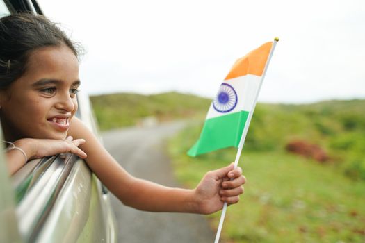 Happy Cheerful Young girl kid holding Indian flag out of in moving car window - Concept of patriotism and Independence or republic day celebration