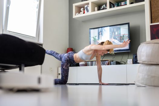 Attractive sporty woman working out at home, doing pilates exercise in front of television in her living room. Social distancing. Stay healthy and stay at home during corona virus pandemic