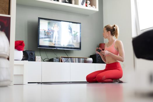 Attractive sporty woman working out at home, doing pilates exercise in front of television in small studio appartment. Social distancing. Stay healthy and stay at home during corona virus pandemic.