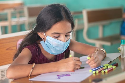 Young girl kid in medical mask busy in drawing sketch at classroom - Concept of safety masures at school due to coronavirus or covid-19 pandemic