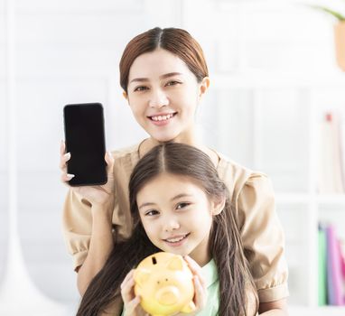 happy mother and daughter showing the mobile phone and piggy bank