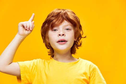 Red-haired boy gestures with his hands in a yellow t-shirt points up with index finger close-up