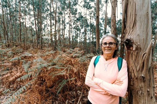 Old woman leaning on a tree while smiling to camera in the forest on sport clothes